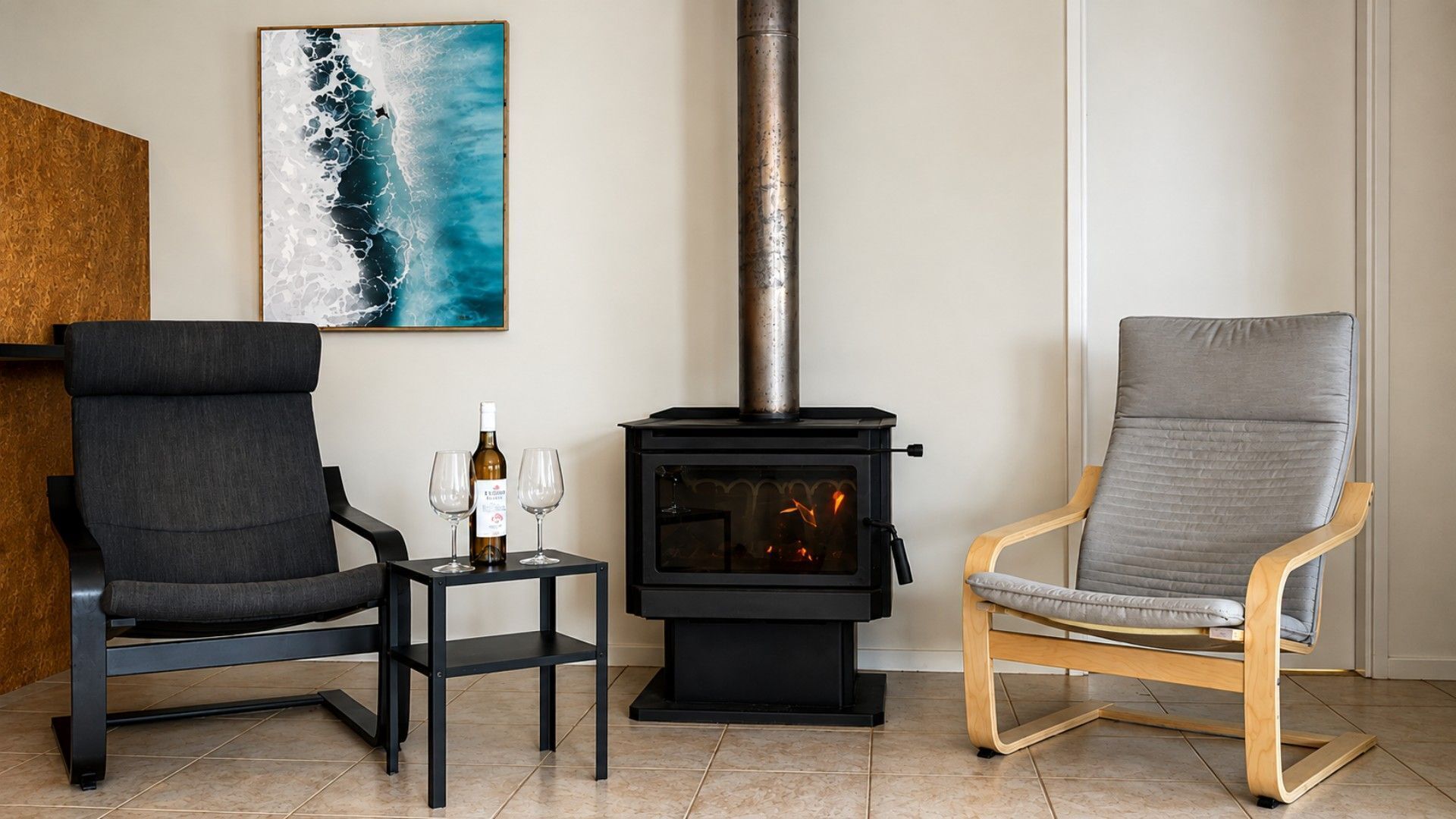 A living room with a black wood-burning stove, two chairs, a small table with wine glasses and a bottle, and a painting of a wave on the wall.