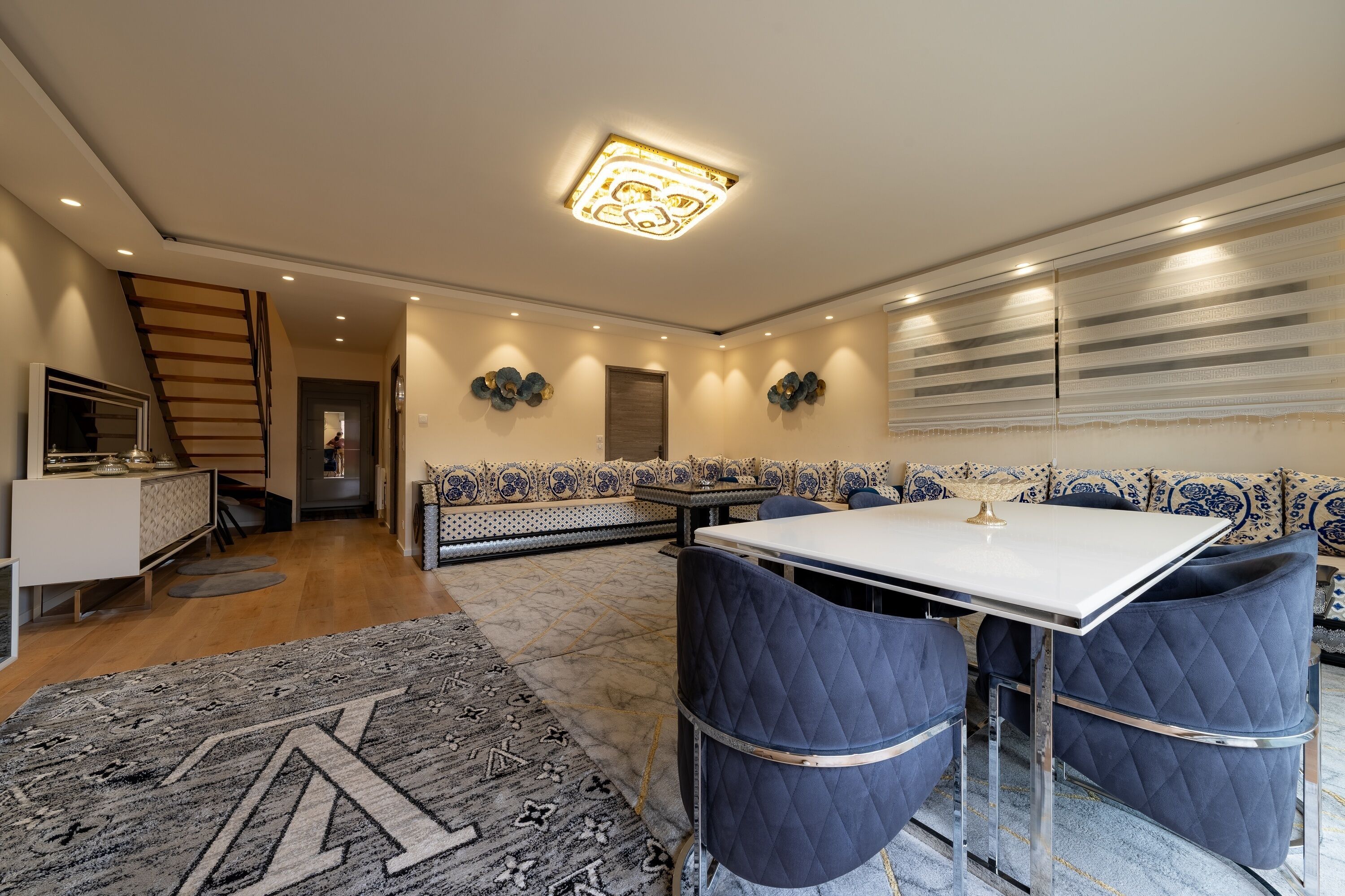 A modern dining area with a white table and blue upholstered chairs, a patterned rug, and decorative wall art.
