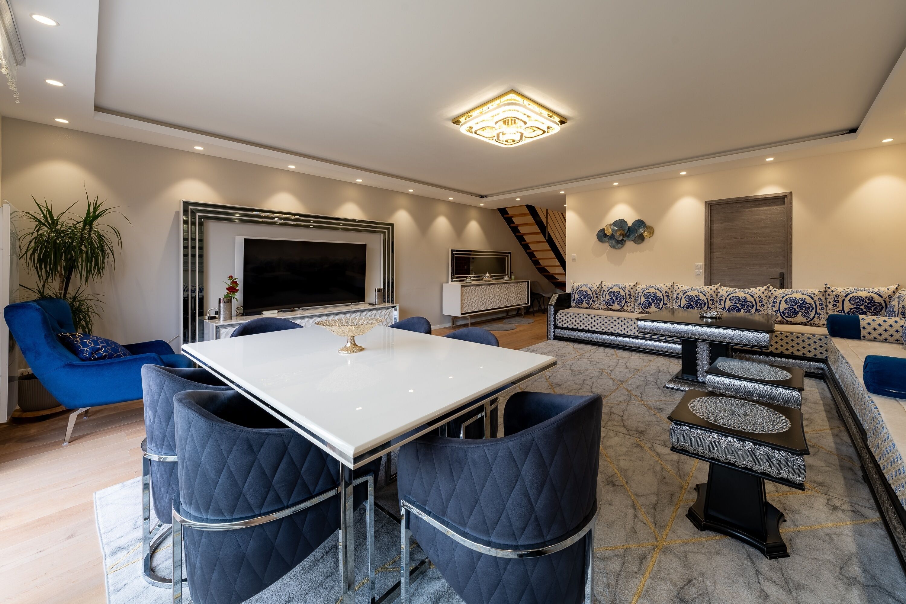 A modern living room with a white dining table, blue chairs, a sofa set, and a staircase.
