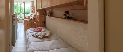 A narrow hallway with a bed, wooden shelves, and a view of an outdoor area.