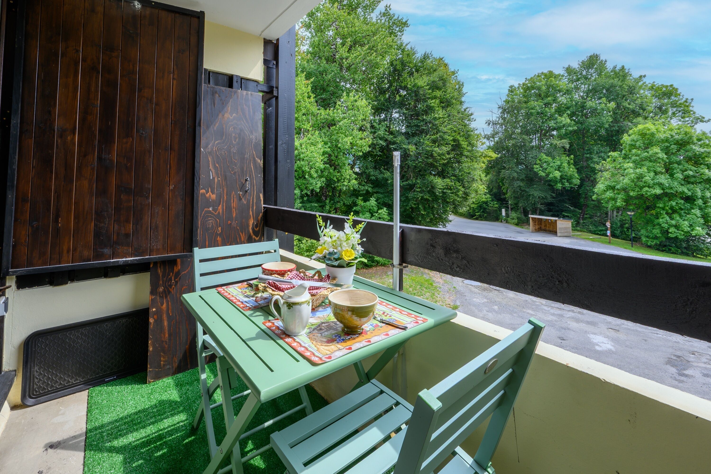Un balcón con una mesa para dos, una puerta de madera y vistas a árboles.