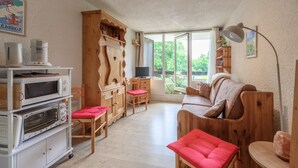 A cozy living room with a wooden sofa, a red chair, a wooden cabinet with kitchen appliances, and a large window offering a view of greenery.