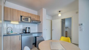 A compact kitchen with wooden cabinets, a microwave, a coffee maker, and a dishwasher. There is a round table with a cutting board and two chairs, one yellow and one white.