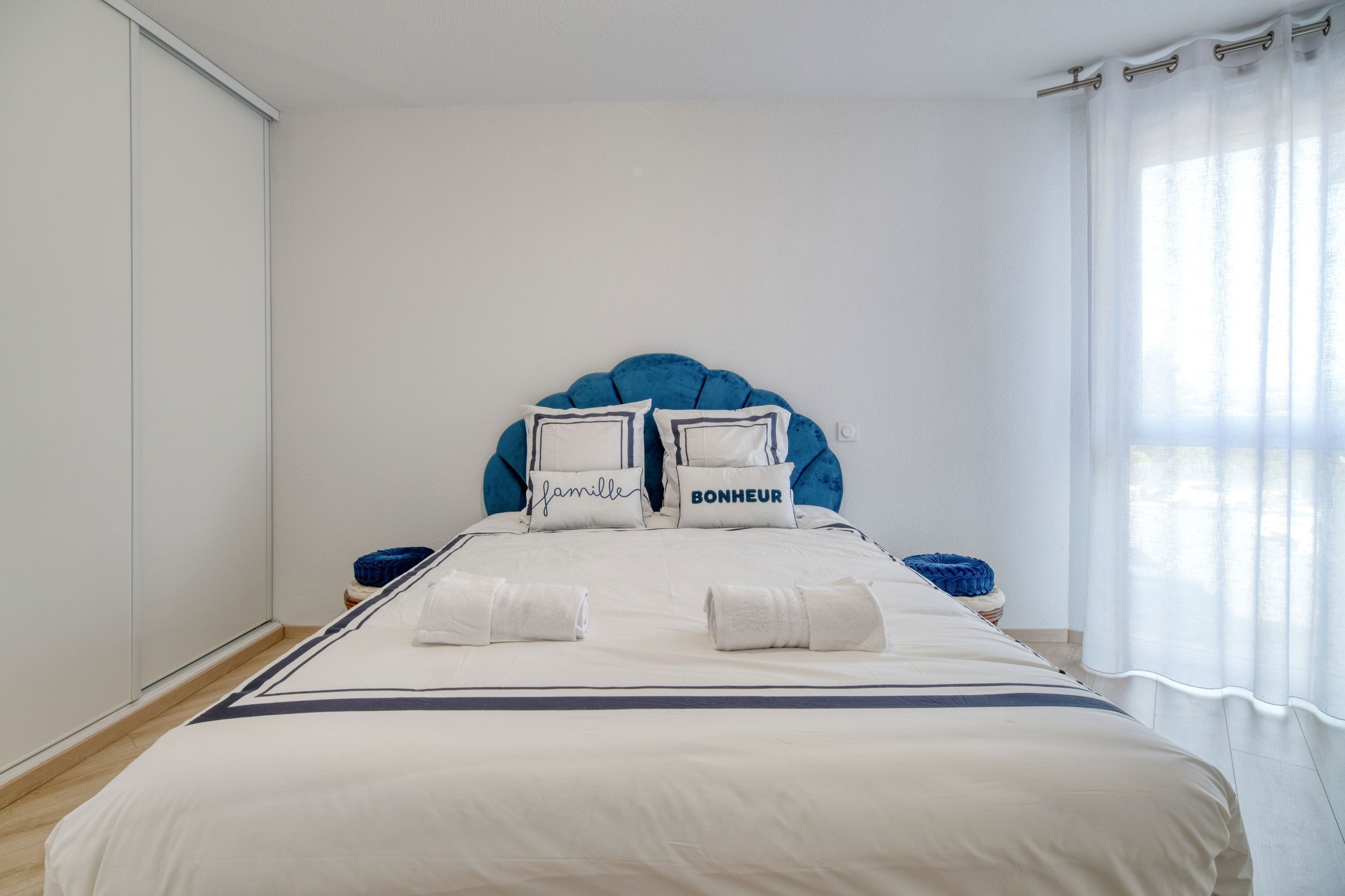 Una cama bien tendida con un cabecero azul, dos almohadas con texto y dos toallas enrolladas.