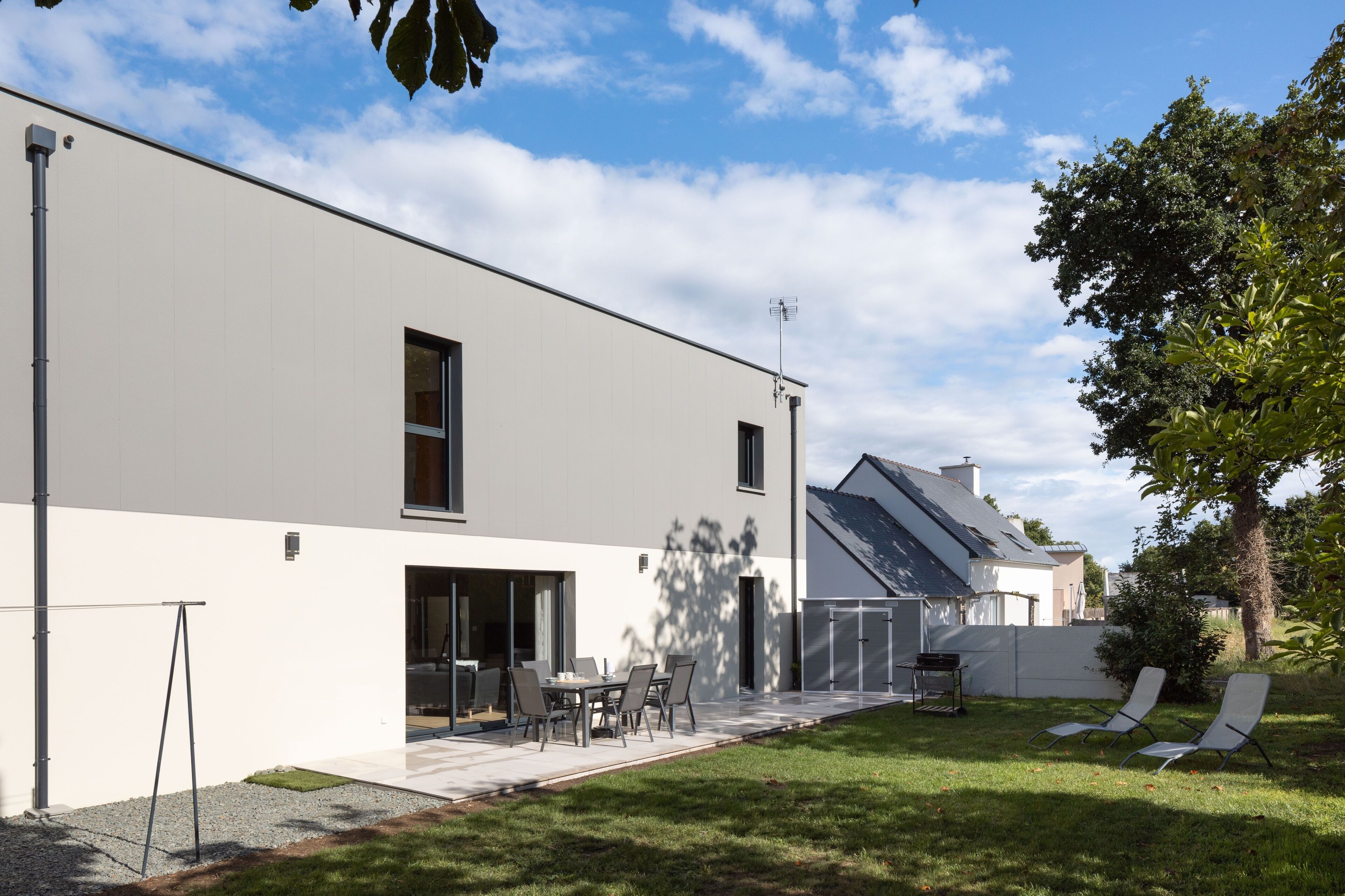 A modern two-story house with a patio, a dining area, and two lounge chairs.