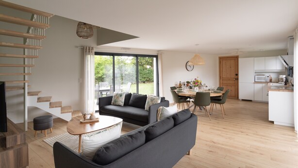 Ein modernes Wohnzimmer mit einer Couch, einem Couchtisch und einem Essbereich mit Stühlen und einem Tisch. Im Hintergrund ist eine Küche mit weißen Schränken und Geräten zu sehen.