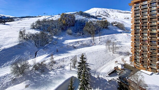 Schneebedeckte Landschaft mit einem modernen Gebäude, Balkonen und einer Seilbahn im Hintergrund.