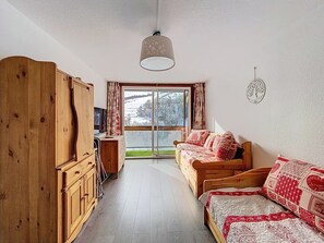 A cozy room with a wooden wardrobe, a sofa with patterned cushions, a television, and a large window with a view of a snowy landscape. - Le Korrigan - Vue Montagne (Villarembert)