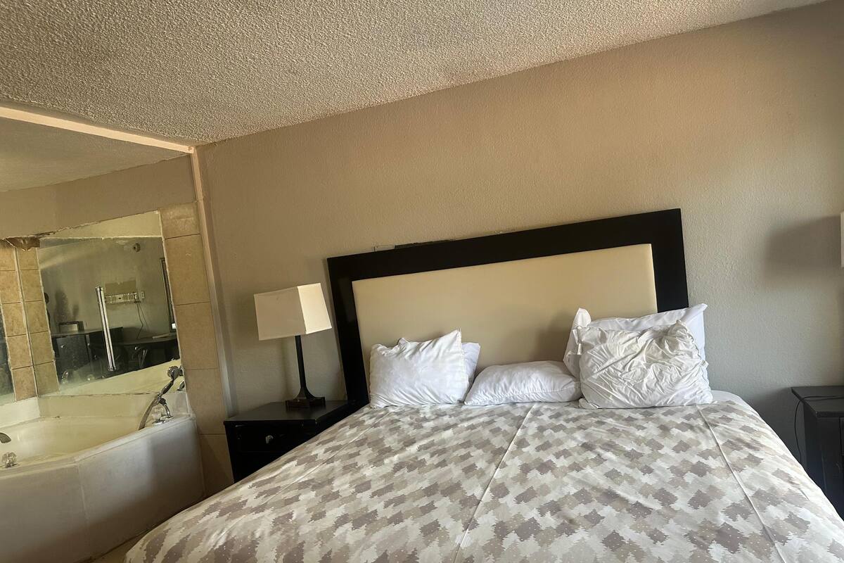 A hotel room with a large bed, a nightstand with a lamp, a bathroom with a bathtub, and a mirror.