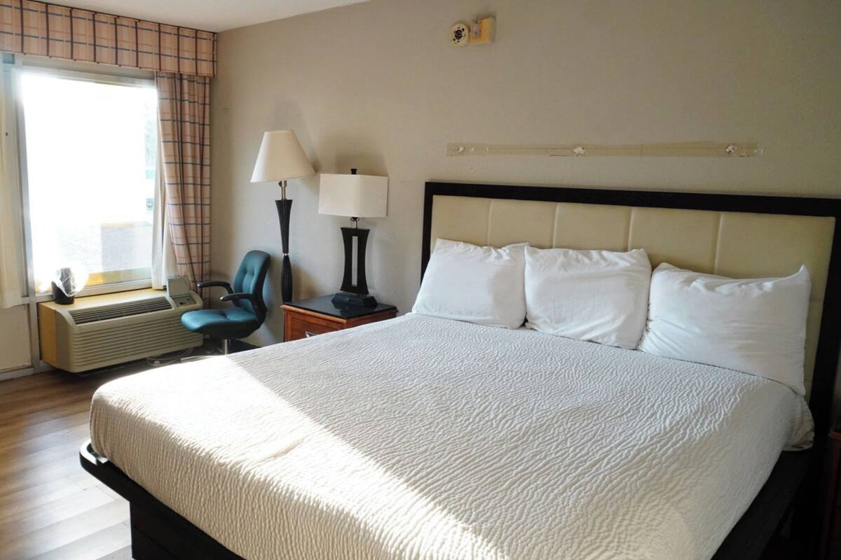 A hotel room with a large bed, a chair, a desk with a lamp, and a window with curtains.