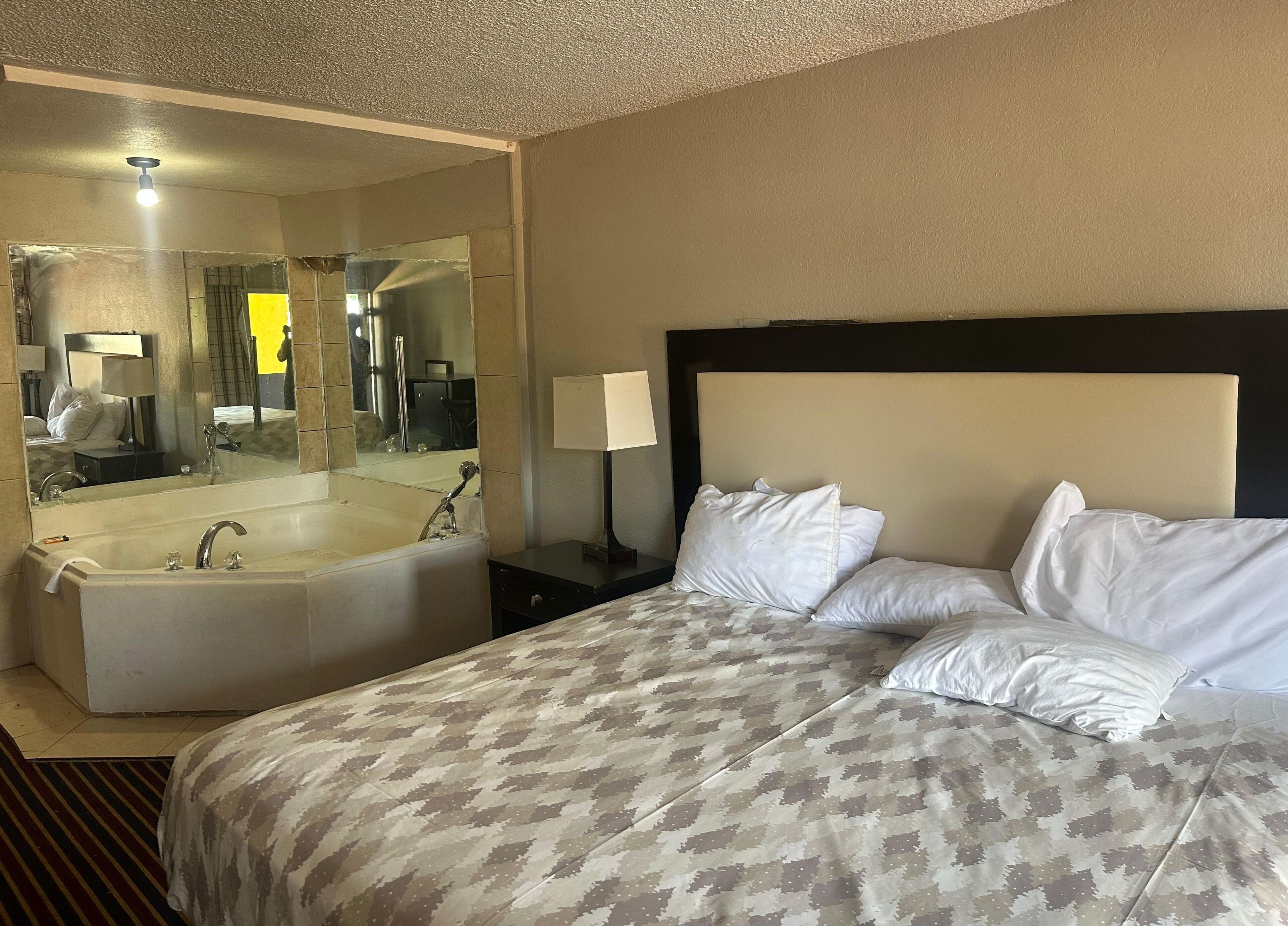 A hotel room with a large bed, a separate bathtub, a nightstand with a lamp, and a mirror reflecting the room.