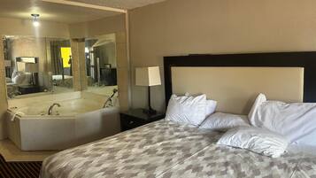 A hotel room with a large bed, a separate bathtub, a nightstand with a lamp, and a mirror reflecting the room.