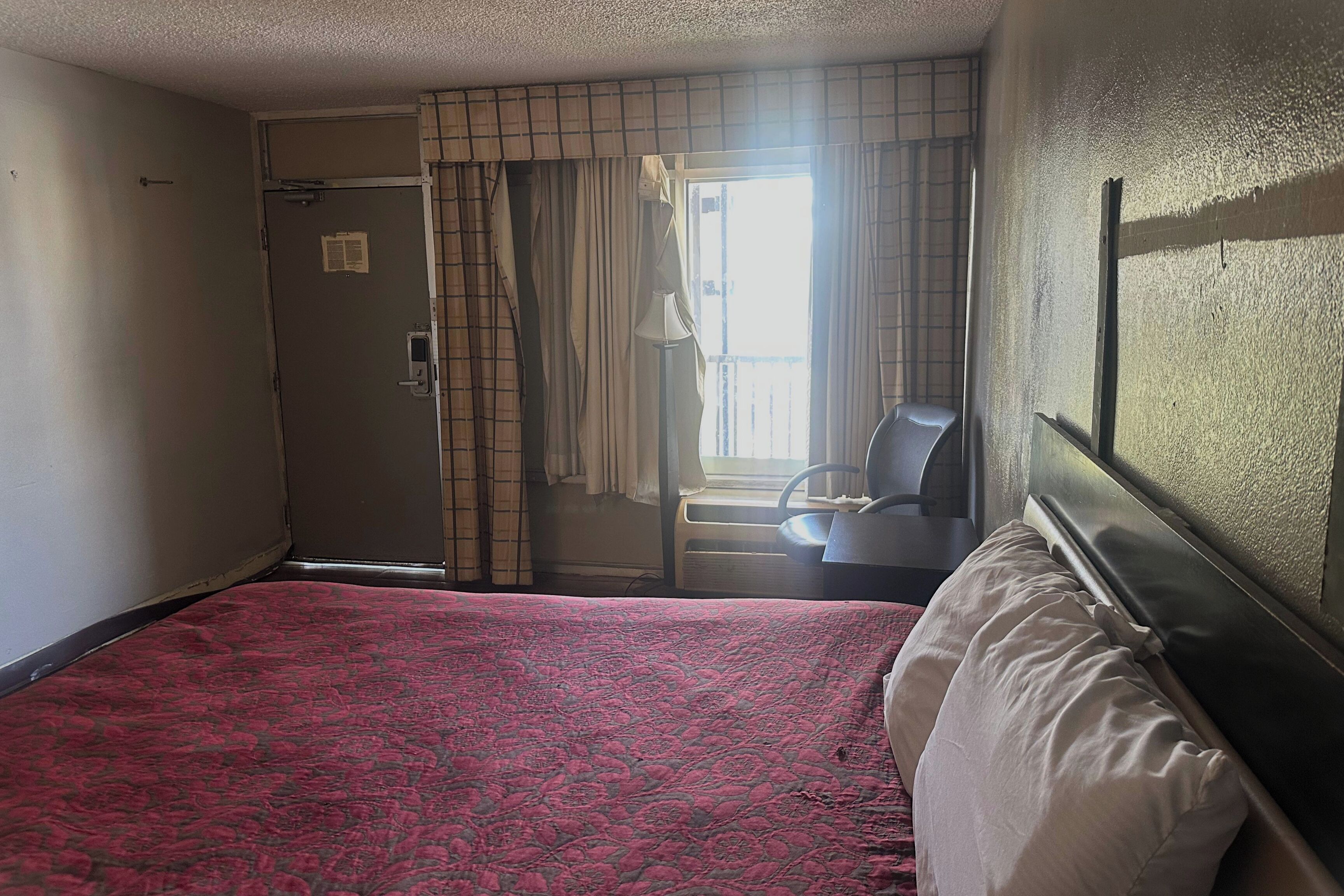 A hotel room with a bed, a chair, a window with curtains, and a door.
