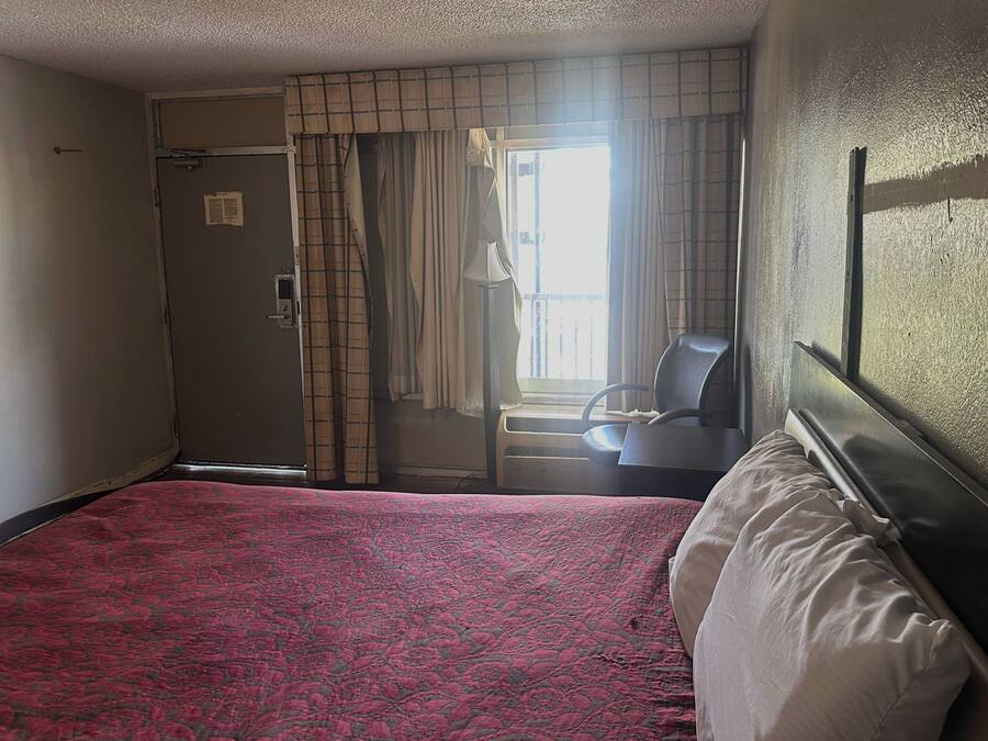 A hotel room with a bed, a chair, a window with curtains, and a door.