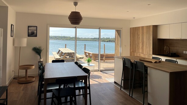 Une cuisine moderne avec des meubles en bois, un coin repas comprenant une table et des chaises, et une vue sur un lac grâce à de grandes portes coulissantes.