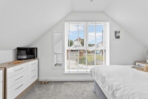 A bedroom with a bed, a television on a dresser, a window with blinds, and a view of a house outside.