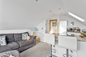 Eine moderne Wohnung mit einem grauen Sofa, einem kleinen Holzschemel, einem weißen Barhocker und einer Küchenzeile mit Toaster und Wasserkocher.