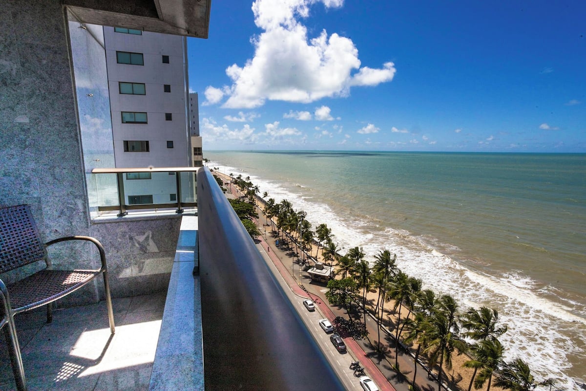 A balcony with a view of the beach, a building, and a parked car.