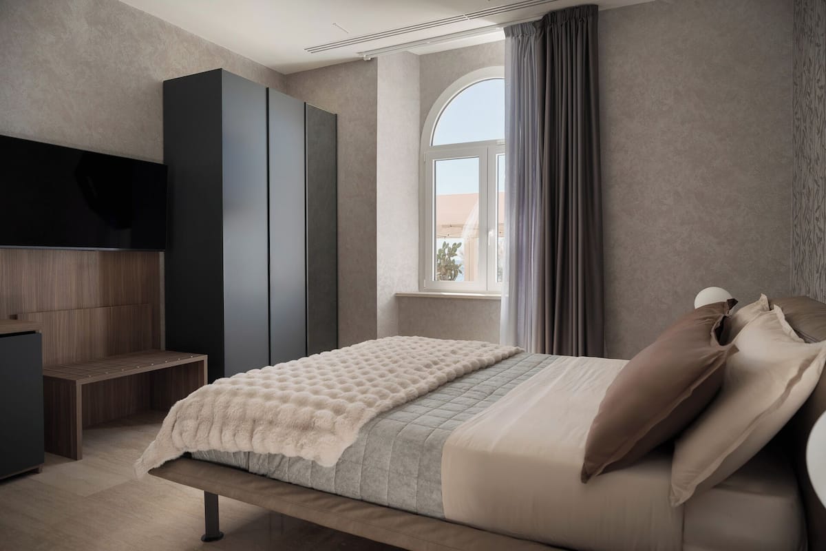 Un dormitorio con cama, armario, banca, televisor y ventana con cortinas.