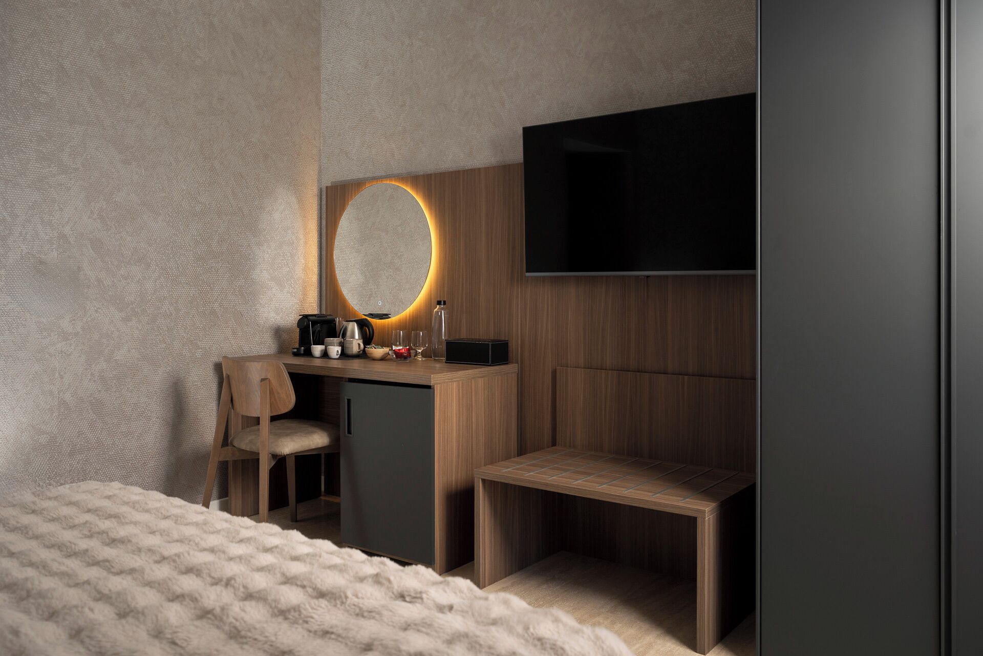 A modern hotel room with a wooden desk, a chair, a small bench, a round mirror with a light, and a flat-screen TV mounted on the wall.