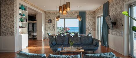 A spacious living room with a large sofa, a coffee table, and a view of the sea through large windows.