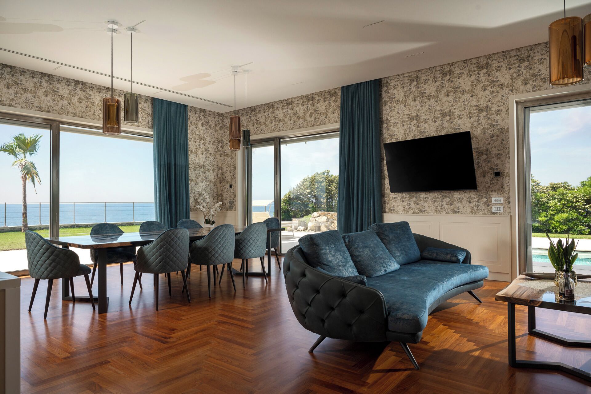 A modern living room with a large sofa, dining table, and chairs, a flat-screen TV, and a view of the ocean.