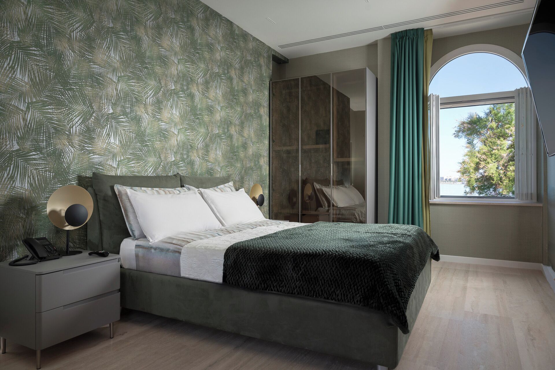 Una camera da letto con un letto, un comodino e vista su un albero attraverso una finestra.