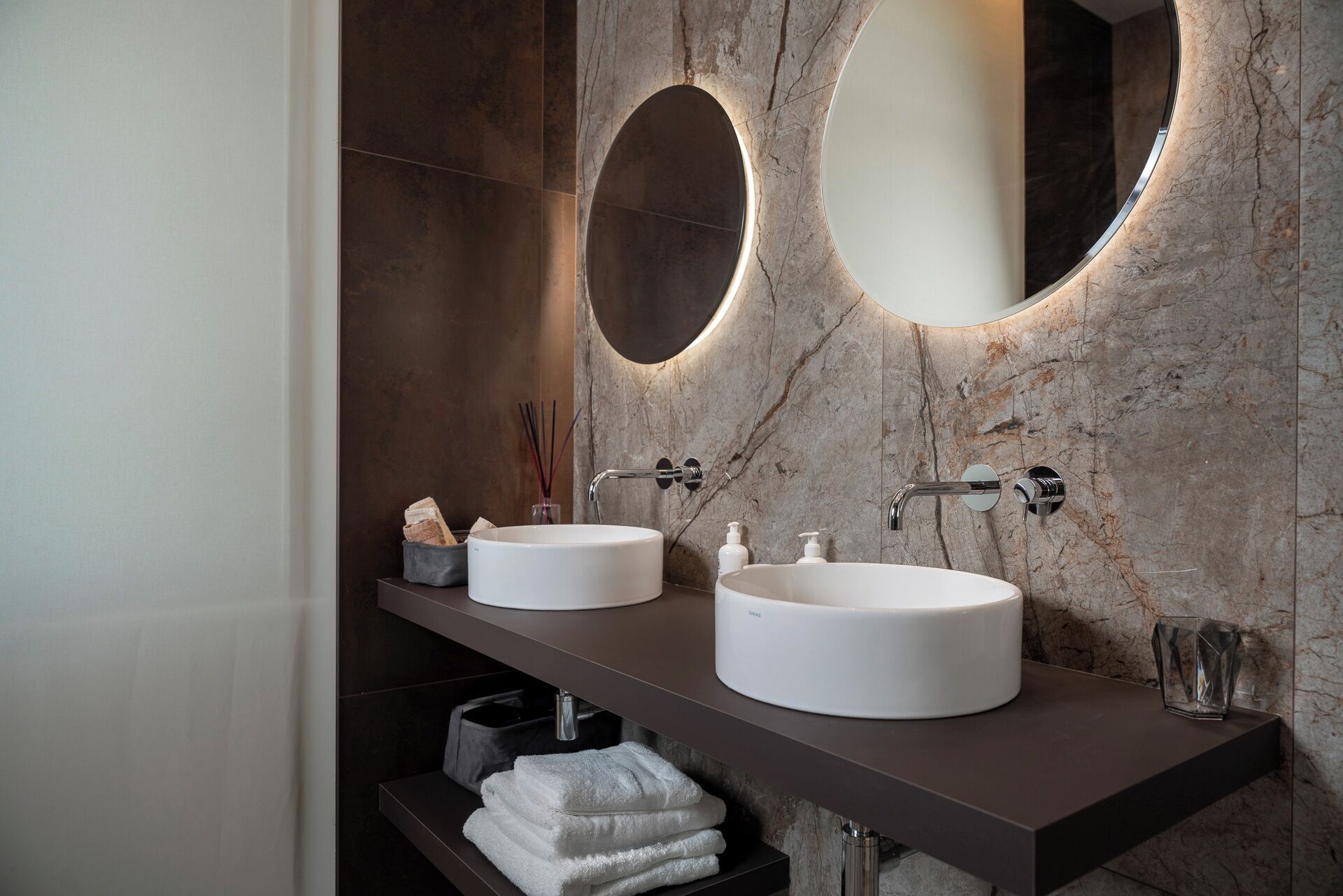 A bathroom with two round white sinks, a dark countertop, and a marble wall.