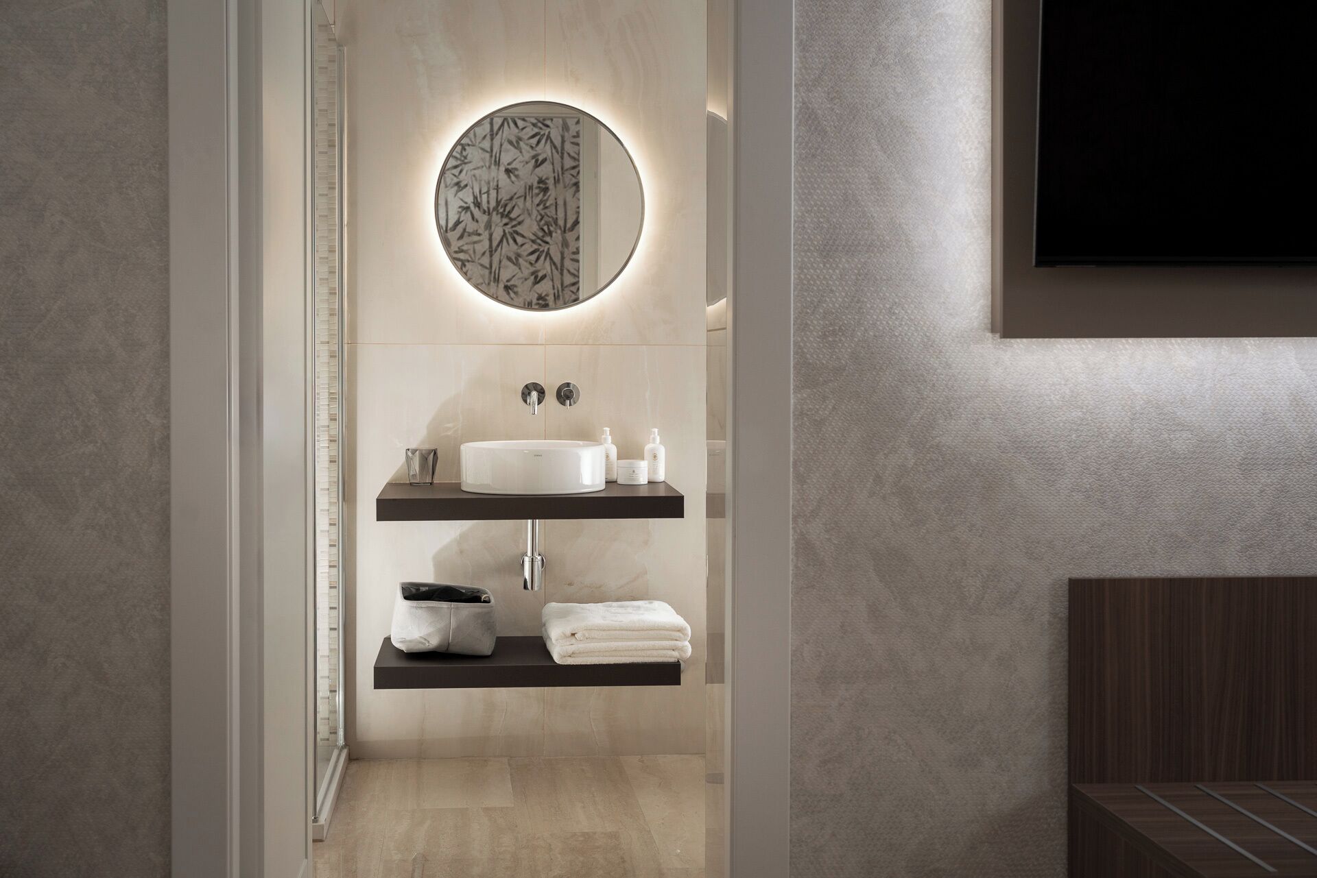 Un baño moderno con un espejo redondo, un lavabo blanco y dos estantes con artículos de aseo.
