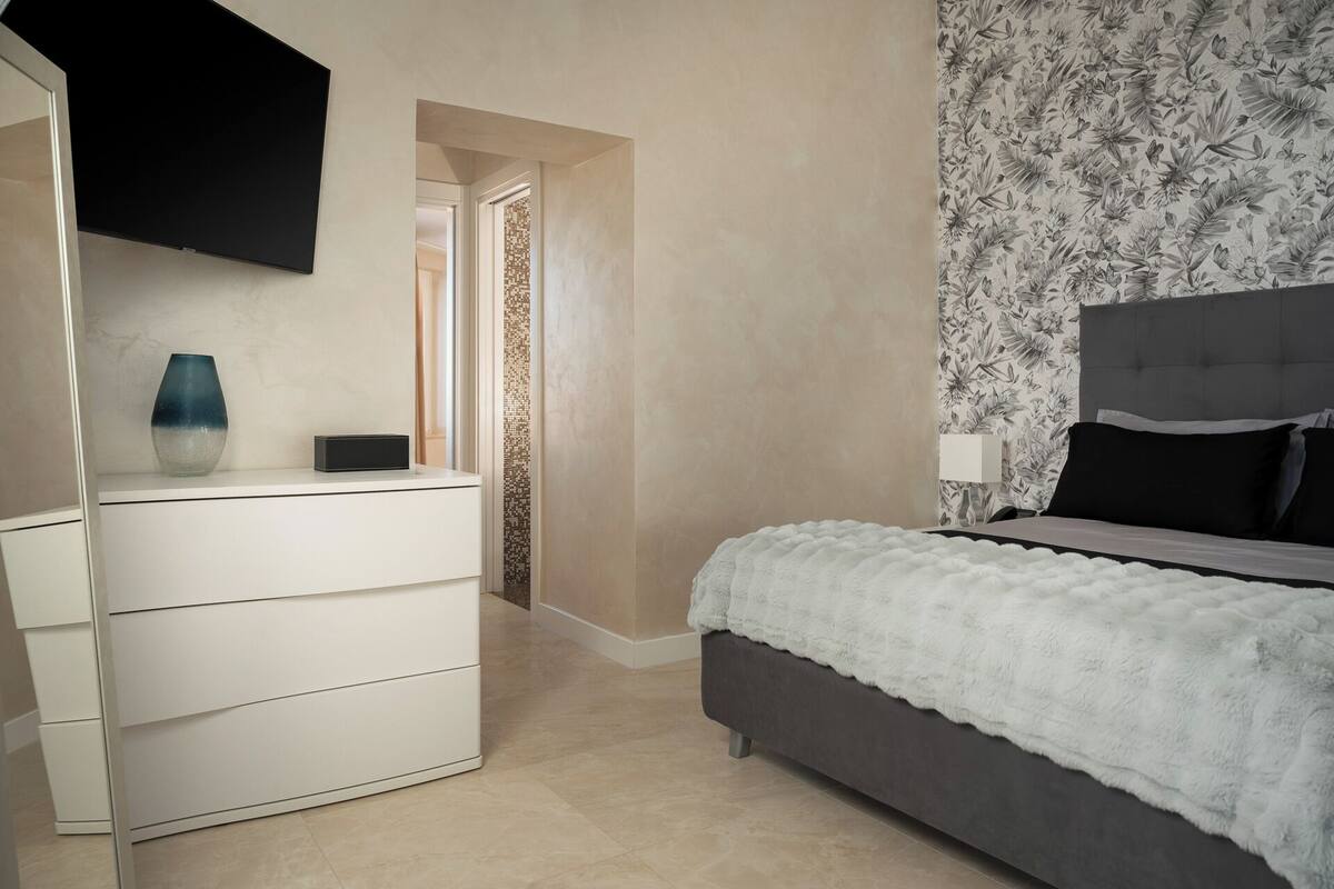 Un dormitorio con cama, cómoda, televisor montado en la pared y un papel tapiz con diseño floral.