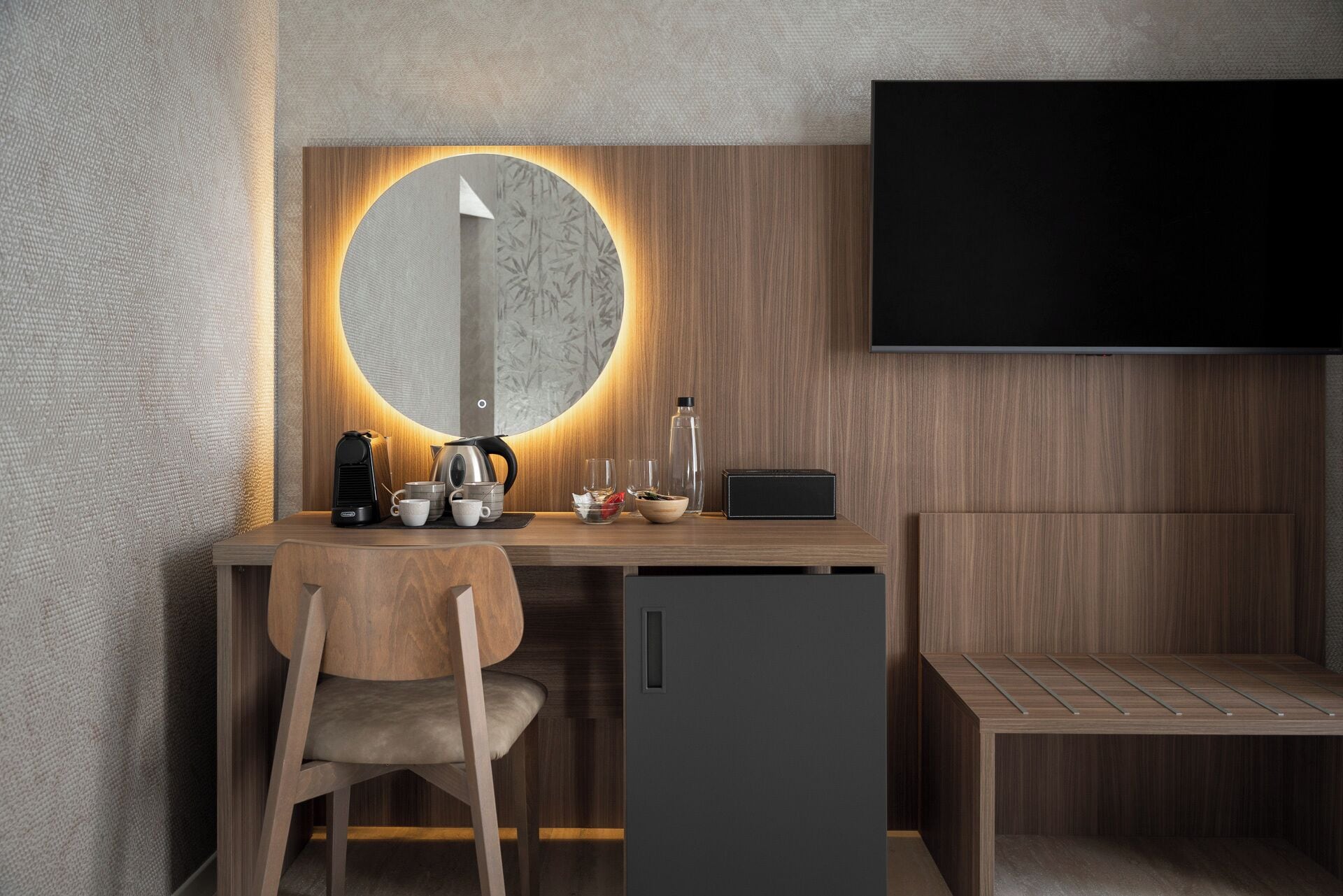 A compact kitchenette with a coffee maker, kettle, and tea set, a small refrigerator, a flat-screen TV, and a wooden chair.
