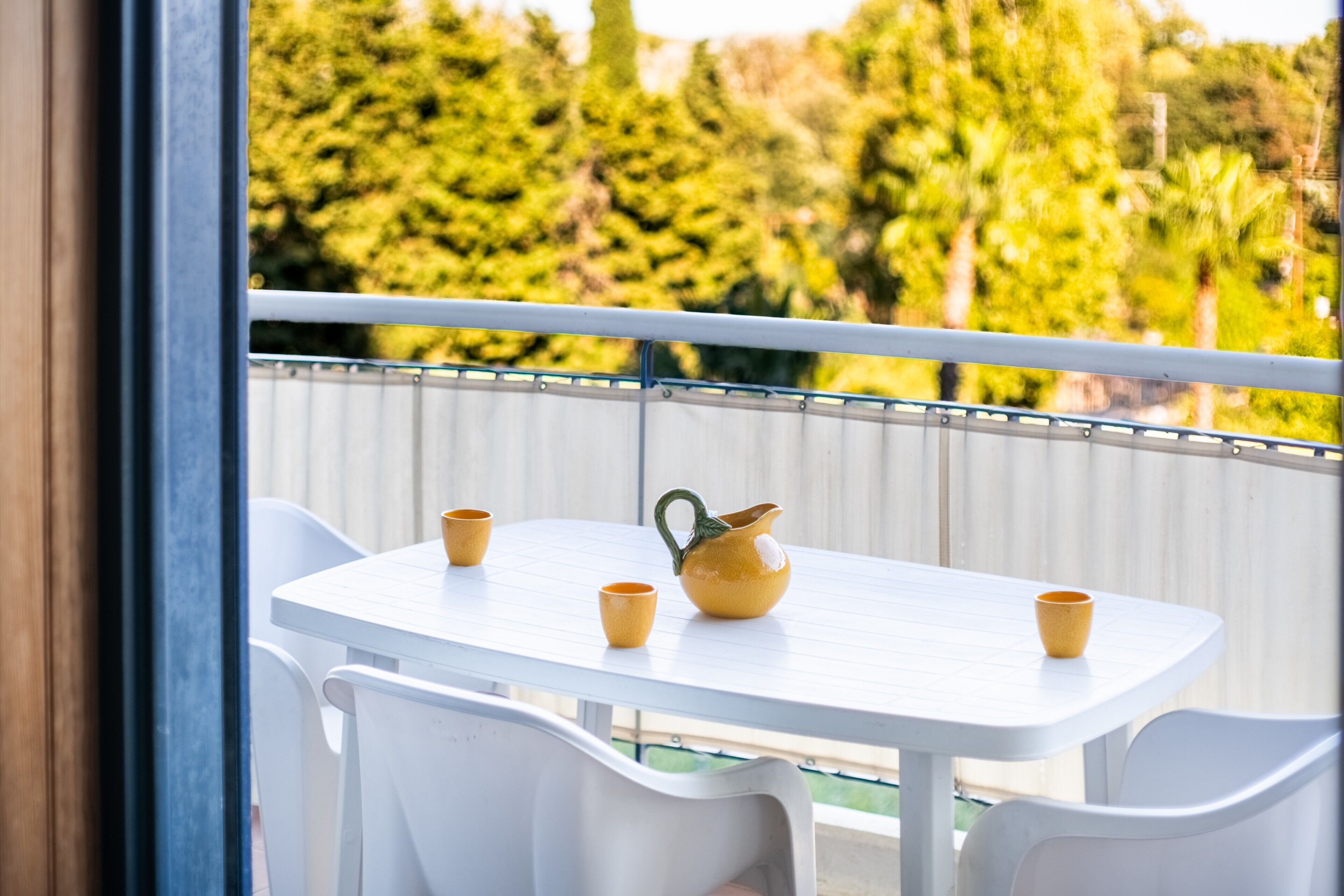 Un balcon avec une table et des chaises blanches, un pichet jaune et quatre tasses jaunes.