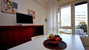 Ein Essbereich mit einem weißen Tisch, einer Obstschale, einem Fernseher an der Wand und einem Fenster mit Vorhängen.