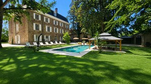 A swimming pool with lounge chairs, a covered seating area, and a large brick building in the background. - Château de la Rochetière (Marcilloles)