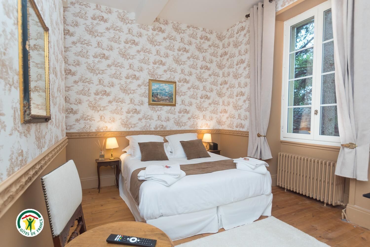 A hotel room with a bed, a nightstand, a chair, a window with curtains, and a radiator.