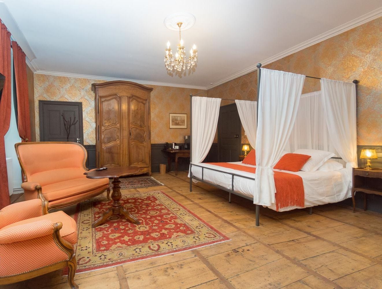 A room with a four-poster bed, a wooden armchair, a television, and a chandelier.