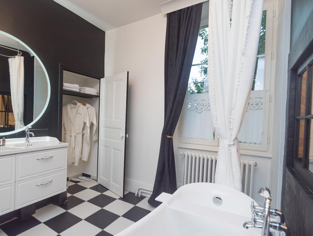 Een moderne badkamer met een zwart-wit schakeerd motief op de vloer, een groot wit bad, een wastafel met spiegel en een inloopkast met opknoping gewaden.
