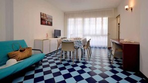 Una habitación con suelo azul y blanco a cuadros, un sofá azul, una mesa de comedor con sillas, un mueble pequeño y un televisor.