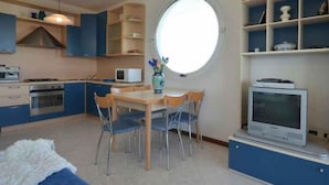 Una cocina compacta con armarios azules, una mesa de comedor con sillas, un ventanal redondo y un televisor sobre un mueble.
