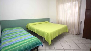 Ein kleines Zimmer mit zwei Einzelbetten, einem grünen Kopfteil, einem gelben Tisch und einem gestreiften Vorhang.
