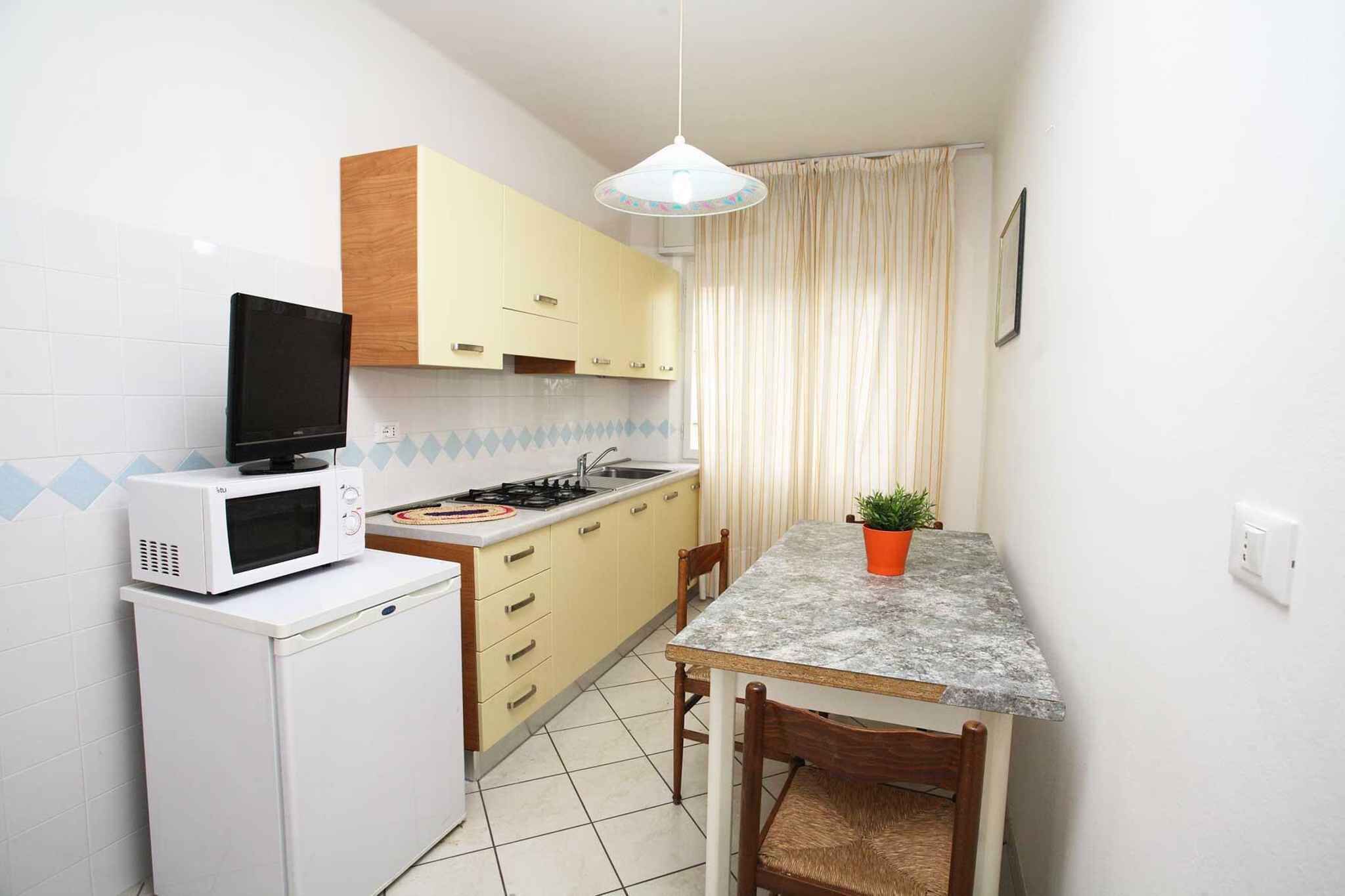 Een praktische keuken met een magnetron, fornuis en spoelbak. Er is een kleine koelkast, een eettafel met stoelen en een potplante.'
