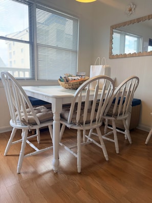 A dining area with a white table and chairs, a basket with items, and a window with blinds.