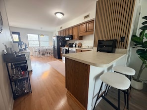 A modern kitchen with wooden cabinets, a central island, bar stools, and a dining area with a table and chairs. - White Marlin & Concerts | 2BR w/ Pool & Parking (Ocean City)