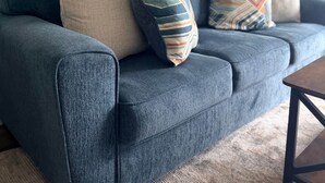 A blue sofa with patterned cushions, a framed artwork on the wall, and a lamp on a side table. - White Marlin & Concerts | 2BR w/ Pool & Parking (Ocean City)