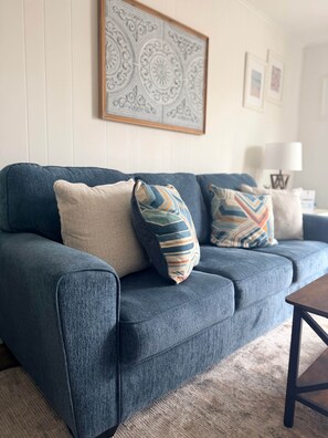 Um sofá azul com almofadas estampadas, uma obra de arte emoldurada na parede e um abajur sobre uma mesinha lateral.