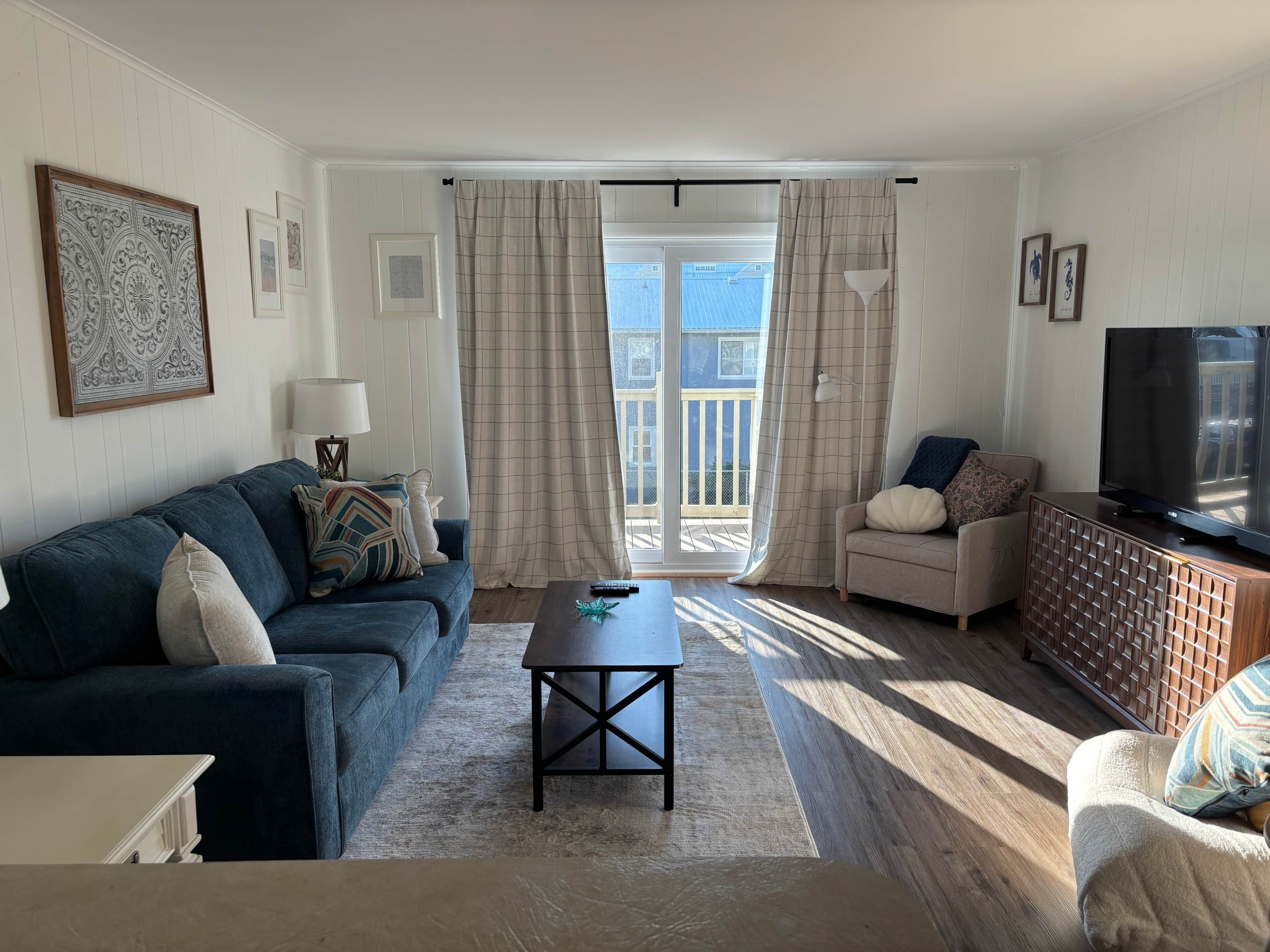 Ein Wohnzimmer mit Sofa, Sessel, Couchtisch, Fernseher und einem Balkon mit Vorhängen.