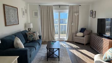 Ein Wohnzimmer mit Sofa, Sessel, Couchtisch, Fernseher und einem Balkon mit Vorhängen.