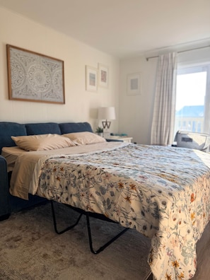 A hotel room with a bed, a nightstand, a lamp, and a window with curtains. - White Marlin & Concerts | 2BR w/ Pool & Parking (Ocean City)