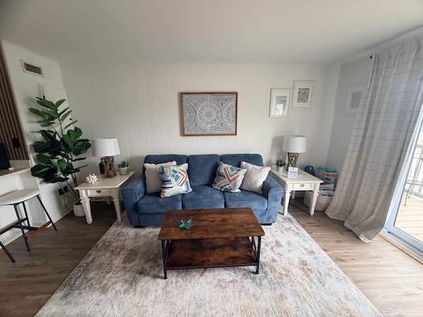 A living room with a blue sofa, a wooden coffee table, a white rug, and a large plant. - White Marlin & Concerts | 2BR w/ Pool & Parking (Ocean City)