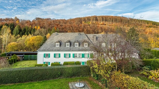 A white building with green shutters, surrounded by a well-maintained garden and a forested hillside. - Apartment Schlossmühle in Bigge-Olsberg (Olsberg)